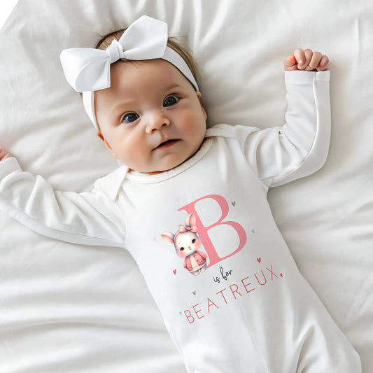 personalised baby outfit: An adorable baby girl wearing a pink sleepsuit for, featuring a cute dusky pink rabbit design. This personalised outfit is available as a sleepsuit, bodysuit, T-shirt, or bib, making it a perfect new baby gift. Ideal for coming home outfits or gender reveal announcements. It has the baby's initial and name along with the pink rabbit design.