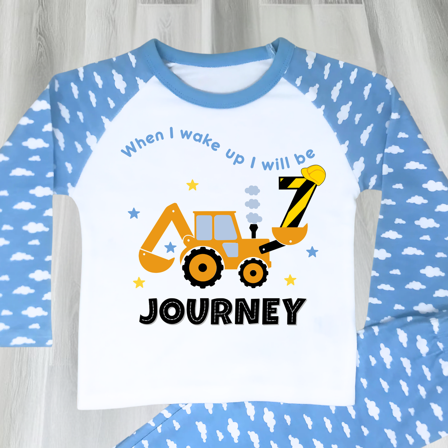 A flat lay blue Pyjama set with a yellow excavator design. The excavator has a black bucket and a black age number on the side. The text 'When I Wake up I will be' is above the excavator and the child's name is below the excavator Digger Construction. Boys personalised birthday gift, Tractor birthday pyjamas, boys custom pyjamas with name, When I wake up construction digger, birthday pyjamas with name
