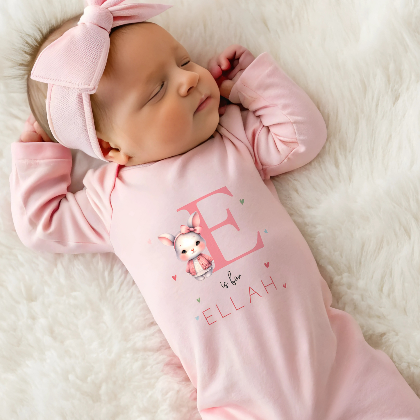 personalised baby outfit: An adorable baby girl wearing a pink sleepsuit for, featuring a cute dusky pink rabbit design. This personalised outfit is available as a sleepsuit, bodysuit, T-shirt, or bib, making it a perfect new baby gift. Ideal for coming home outfits or gender reveal announcements. It has the baby's initial and name along with the pink rabbit design.