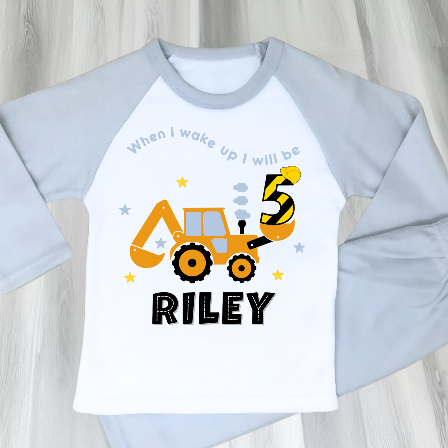 A flat lay blue Pyjama set with a yellow excavator design. The excavator has a black bucket and a black age number on the side. The text 'When I Wake up I will be' is above the excavator and the child's name is below the excavator Digger Construction. Boys personalised birthday gift, Tractor birthday pyjamas, boys custom pyjamas with name, When I wake up construction digger, birthday pyjamas with name