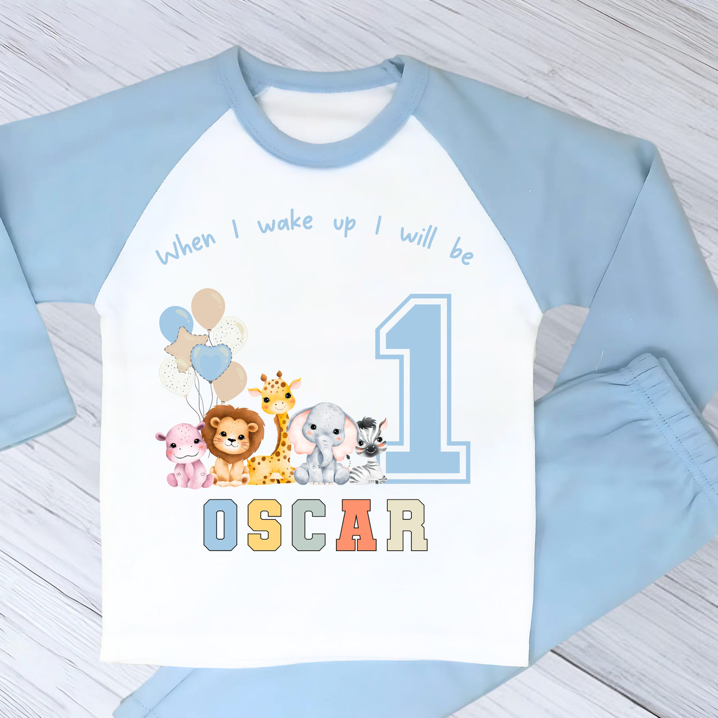 Safari Birthday pyjamas, A personalised set of boys birthday pyjamas in blue. At the top it says when i wake up i will be followed by some safari jungle animals holding some birthday balloons. They are sat beside a number that represents the child's age on birthday. Under that is the child name in safari rainbow colours. This is perfect gift for a toddler first birthday.