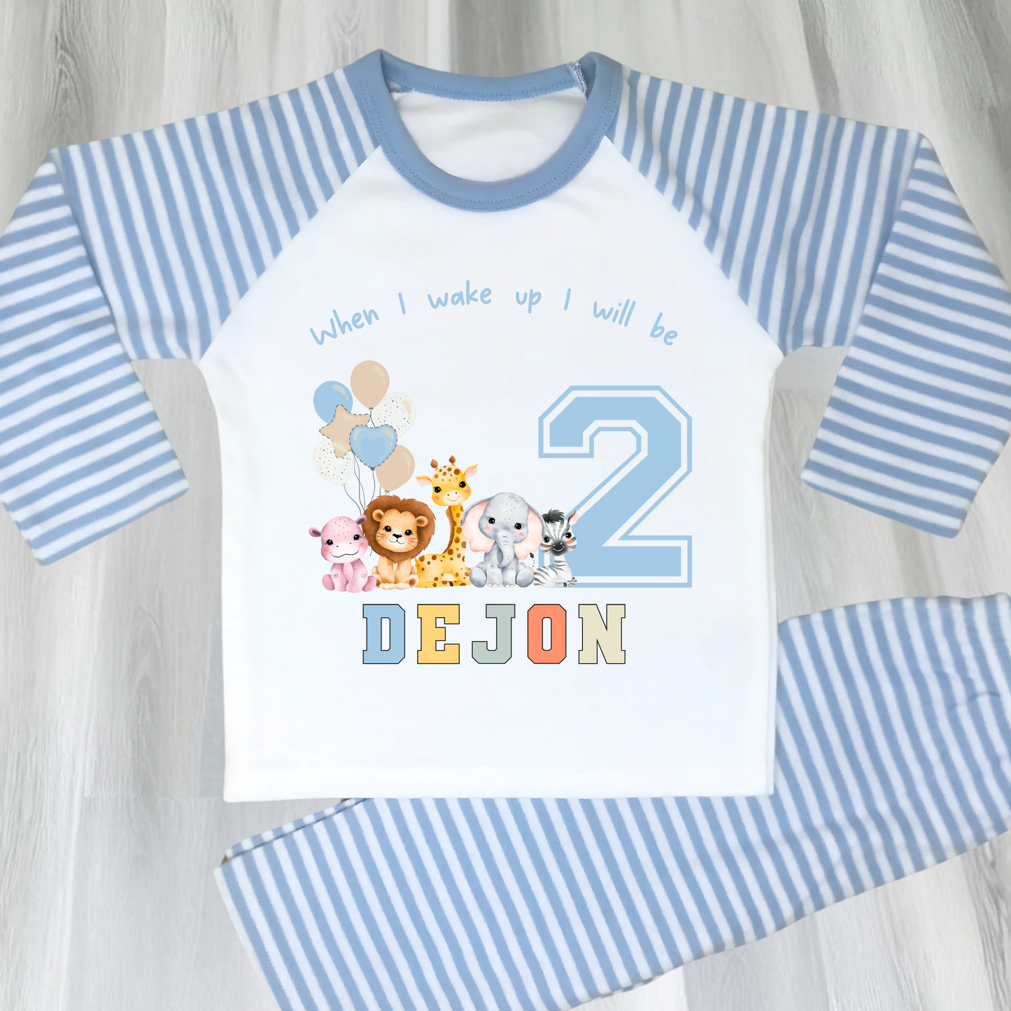 Safari Birthday pyjamas, A personalised set of boys birthday pyjamas in blue. At the top it says when i wake up i will be followed by some safari jungle animals holding some birthday balloons. They are sat beside a number that represents the child's age on birthday. Under that is the child name in safari rainbow colours. This is perfect gift for a toddler first birthday.