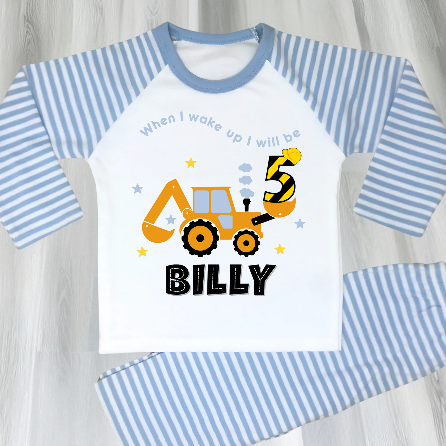 A flat lay blue Pyjama set with a yellow excavator design. The excavator has a black bucket and a black age number on the side. The text 'When I Wake up I will be' is above the excavator and the child's name is below the excavator Digger Construction. Boys personalised birthday gift, Tractor birthday pyjamas, boys custom pyjamas with name, When I wake up construction digger, birthday pyjamas with name