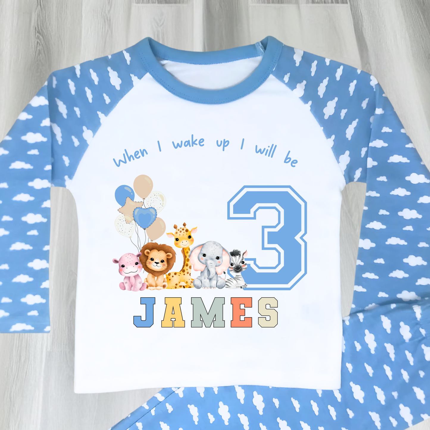 Safari Birthday pyjamas, A personalised set of boys birthday pyjamas in blue. At the top it says when i wake up i will be followed by some safari jungle animals holding some birthday balloons. They are sat beside a number that represents the child's age on birthday. Under that is the child name in safari rainbow colours. This is perfect gift for a toddler first birthday.