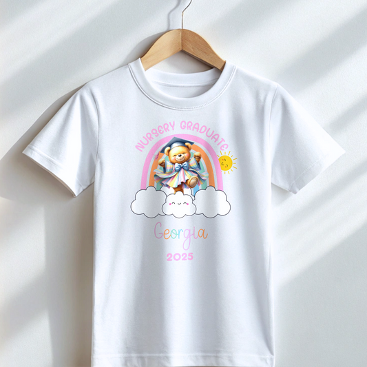 A girl wearing a white Personalised Nursery Leavers T-shirt. Above the rainbow it says Nursery Graduate and below it is a girl bear in graduation clothes on soft fluffy clouds. Below the design is the girls name and year of graduation.