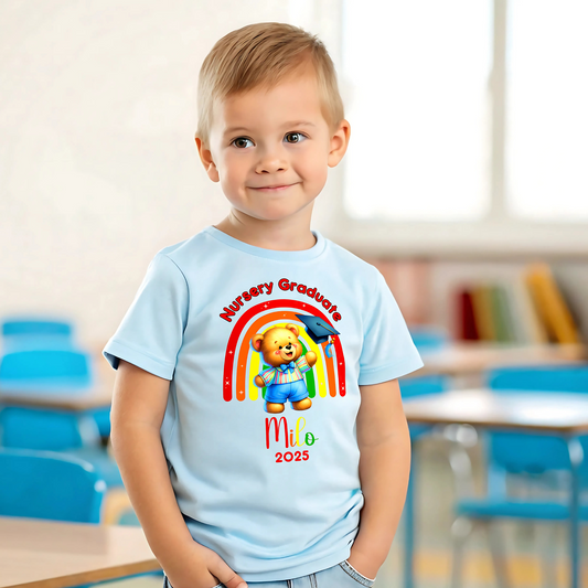 A girl wearing a white Personalised Nursery Leavers T-shirt. Above the rainbow it says Nursery Graduate and below it is a girl bear in graduation clothes on soft fluffy clouds. Below the design is the girls name and year of graduation.