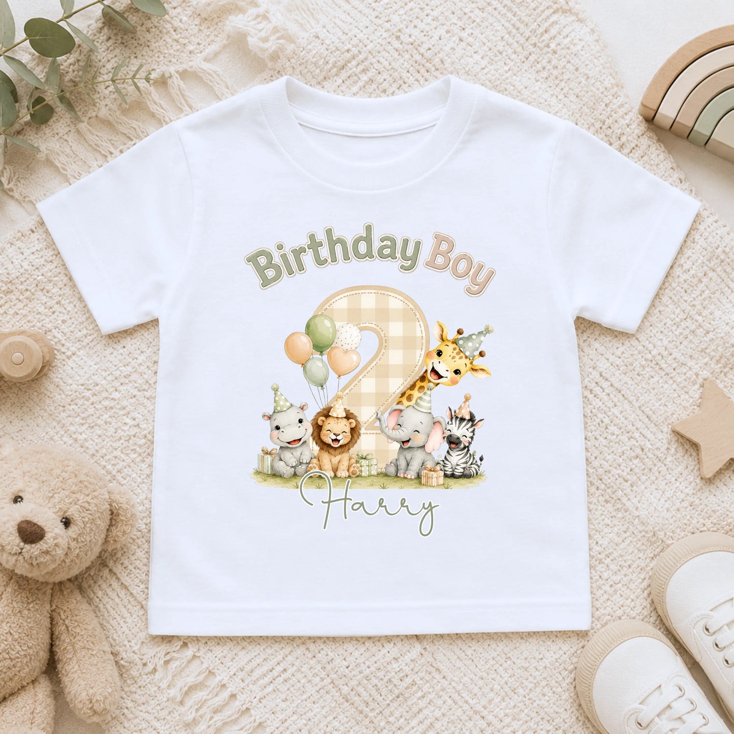 A white short-sleeve baby t-shirt with a colorful 'Birthday Boy Harry' animal print, laid flat on a beige textured blanket surrounded by toys.