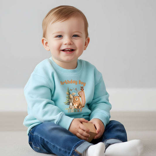 Woodland animal birthday sweatshirt: A children's birthday jumper featuring a woodland animal theme. The shirt displays a forest scene with a playful fox, a curious bear, and a gentle bear, surrounded by lush greenery. The design uses earthy tones and vibrant greens to create a natural look. At the top, the words “Birthday Boy” are printed, with space for a personalized name underneath.