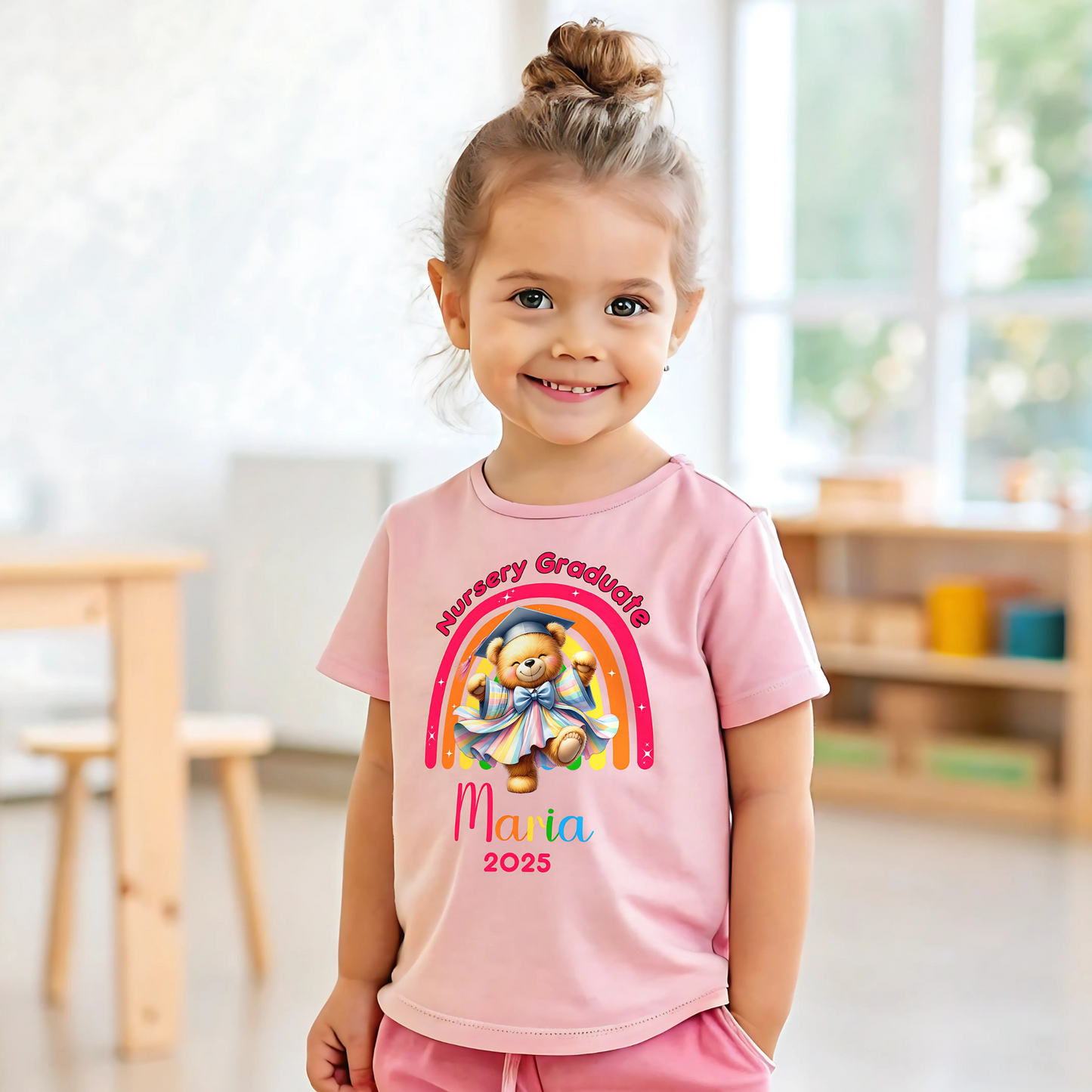 A girl wearing a white Personalised Nursery Leavers T-shirt. Above the rainbow it says Nursery Graduate and below it is a girl bear in graduation clothes on soft fluffy clouds. Below the design is the girls name and year of graduation.