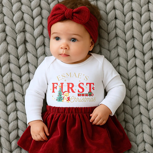 First Chrstmas Outfit, Baby wearing a white long-sleeve shirt with personalised First Christmas' text and a red skirt, lying on a textured gray blanket.