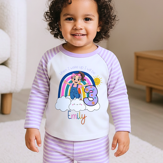 Personalised Ms Rachel Birthday PJs for Girls, When I Wake up Ms Rachel Pyjamas AishaBaileyDesigns: A smiling toddler wearing a custom pajama set with lavender and white stripes, featuring a white front panel with a colorful cartoon rainbow, character, the number 3, and the name 'Emily'.