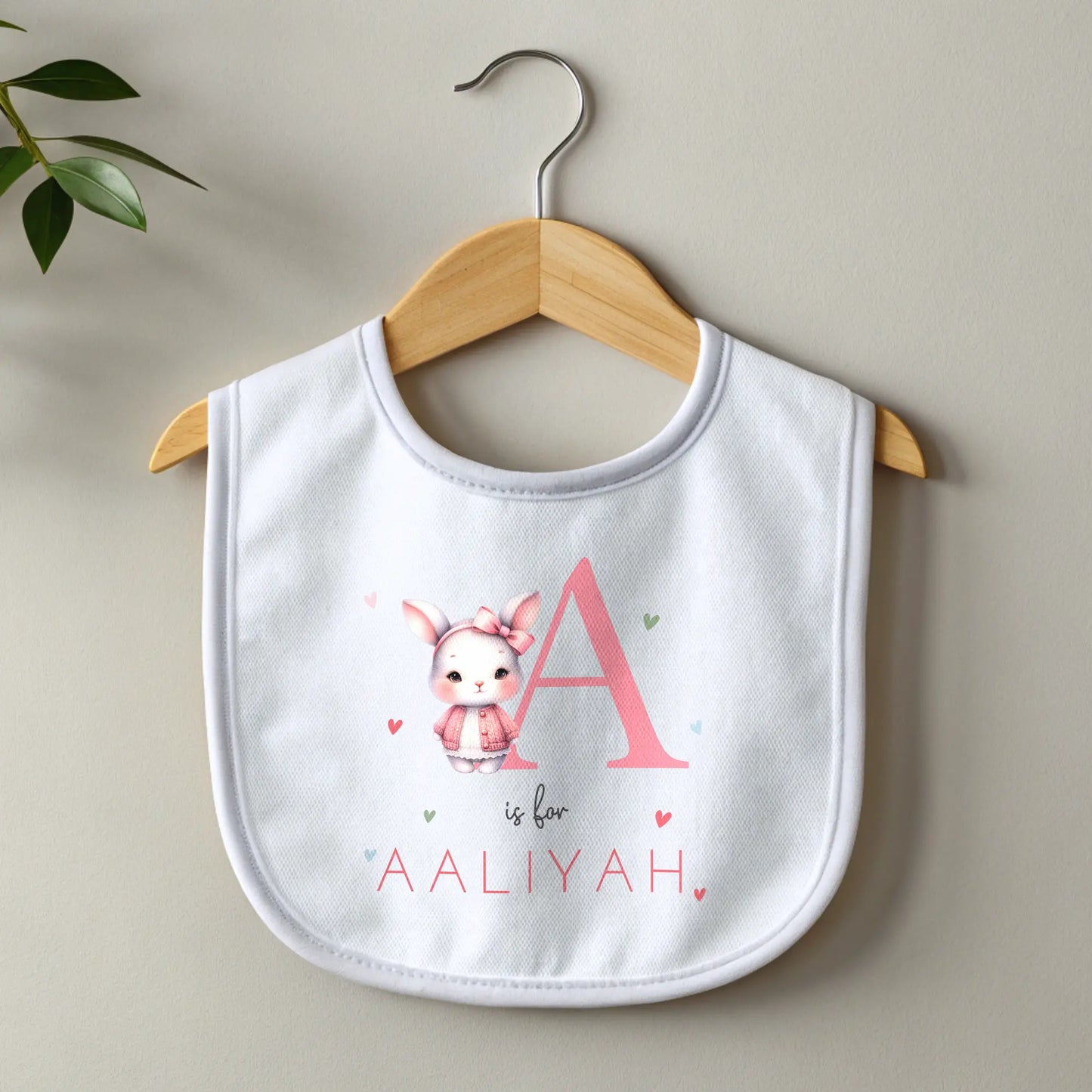 personalised baby outfit: An adorable baby girl wearing a pink sleepsuit for, featuring a cute dusky pink rabbit design. This personalised outfit is available as a sleepsuit, bodysuit, T-shirt, or bib, making it a perfect new baby gift. Ideal for coming home outfits or gender reveal announcements. It has the baby's initial and name along with the pink rabbit design.