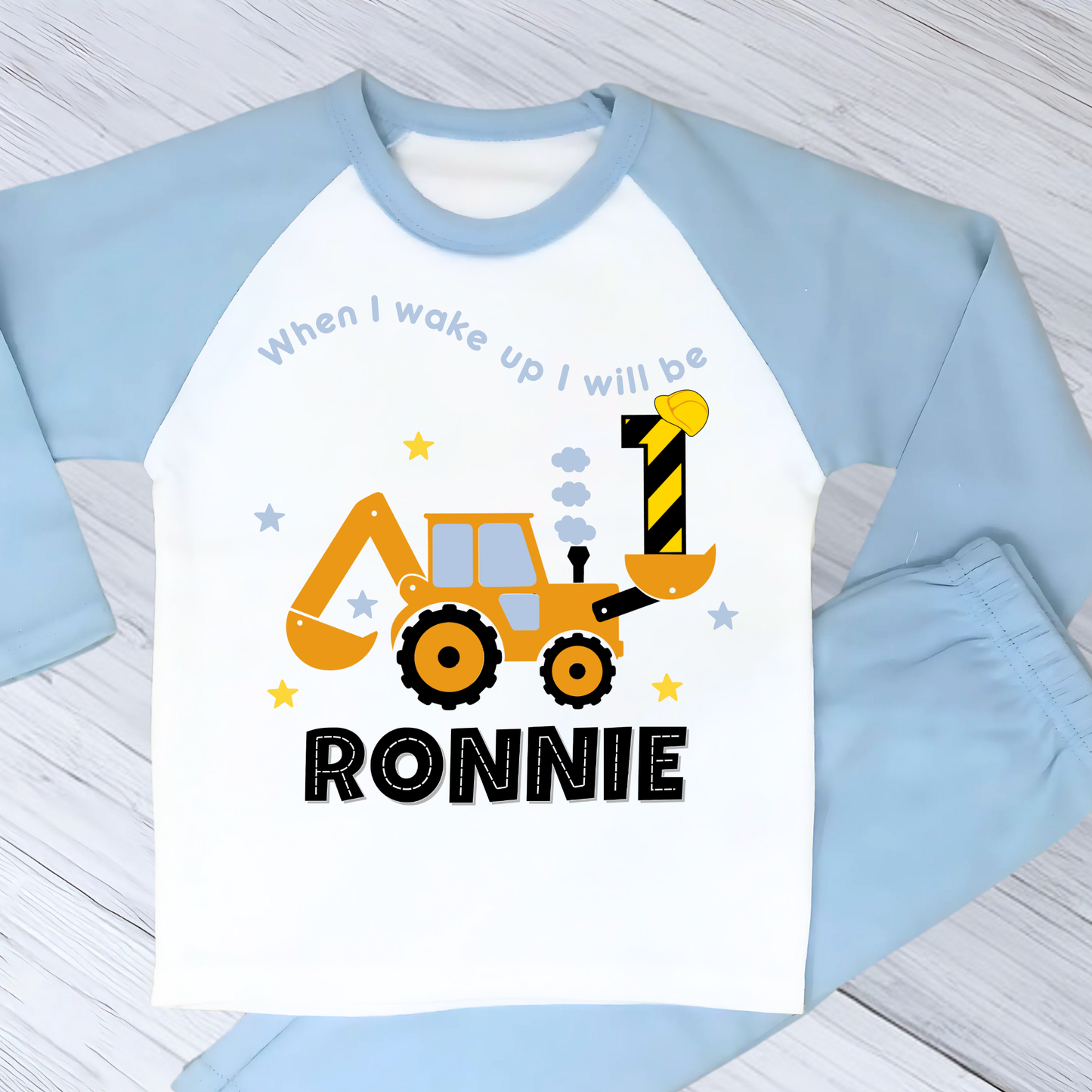 A flat lay blue Pyjama set with a yellow excavator design. The excavator has a black bucket and a black age number on the side. The text 'When I Wake up I will be' is above the excavator and the child's name is below the excavator Digger Construction. Boys personalised birthday gift, Tractor birthday pyjamas, boys custom pyjamas with name, When I wake up construction digger, birthday pyjamas with name