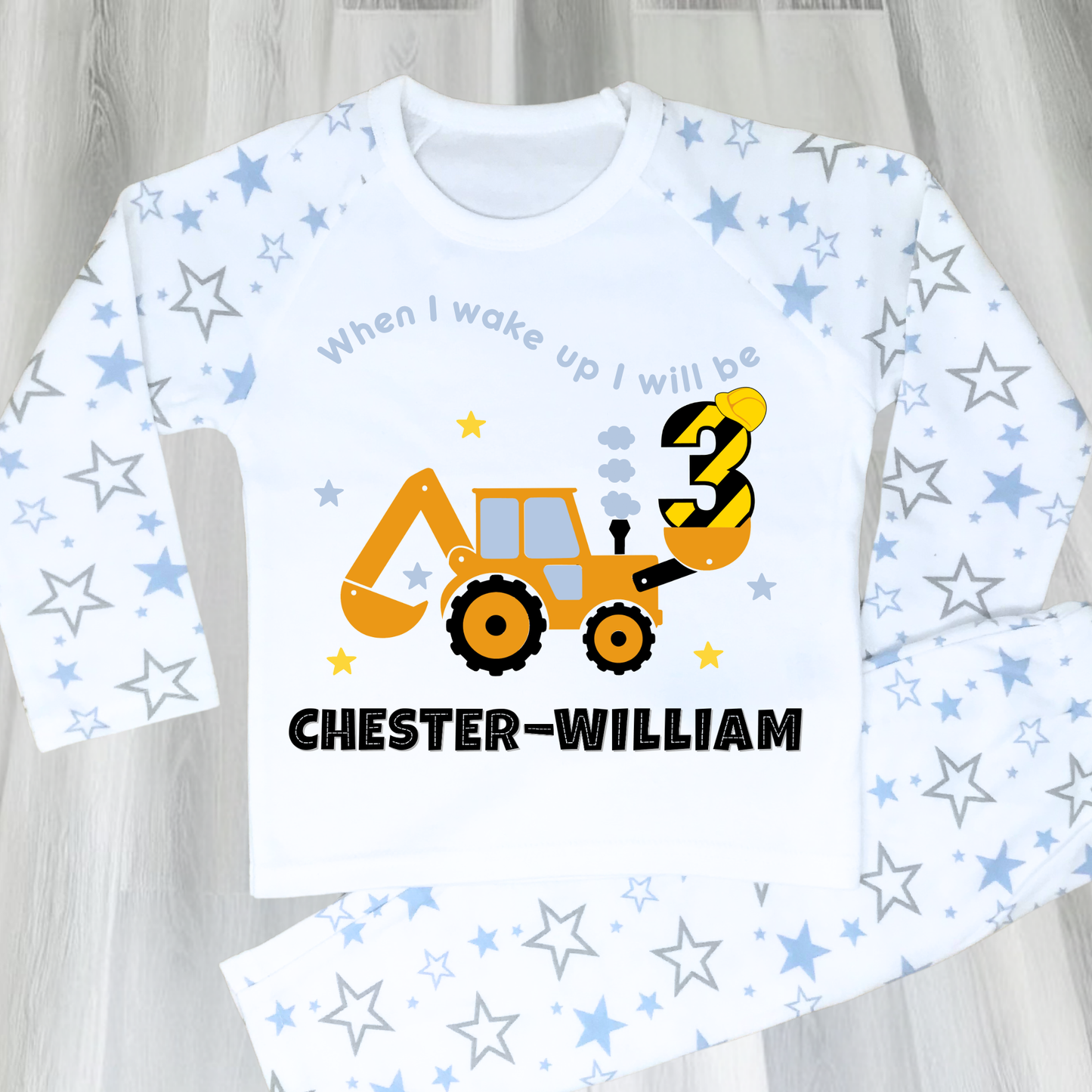 A flat lay blue Pyjama set with a yellow excavator design. The excavator has a black bucket and a black age number on the side. The text 'When I Wake up I will be' is above the excavator and the child's name is below the excavator Digger Construction. Boys personalised birthday gift, Tractor birthday pyjamas, boys custom pyjamas with name, When I wake up construction digger, birthday pyjamas with name