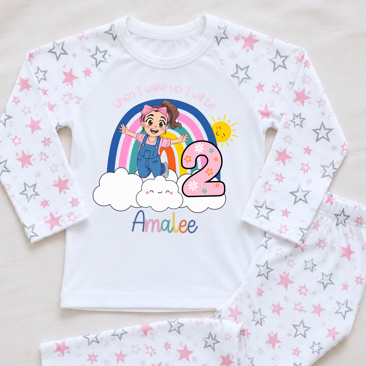 Personalised Ms Rachel Birthday PJs for Girls, When I Wake up Ms Rachel Pyjamas AishaBaileyDesigns: A two-piece children's pajama set laid flat on a light beige surface. The long-sleeve top is white with an all-over pink and gray star pattern, featuring a colorful cartoon birthday graphic with the text 'When I wake up I will be 2 Amalee'.