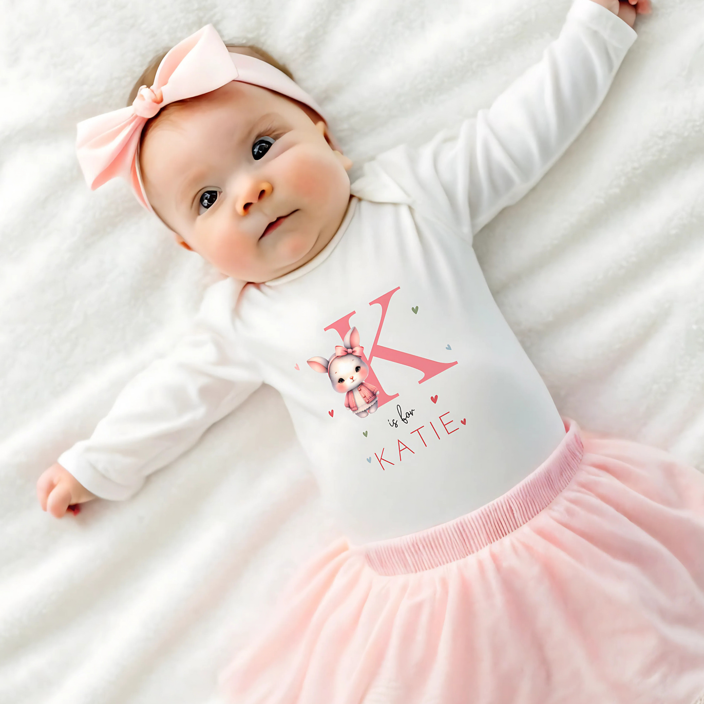 personalised baby outfit: An adorable baby girl wearing a pink sleepsuit for, featuring a cute dusky pink rabbit design. This personalised outfit is available as a sleepsuit, bodysuit, T-shirt, or bib, making it a perfect new baby gift. Ideal for coming home outfits or gender reveal announcements. It has the baby's initial and name along with the pink rabbit design.