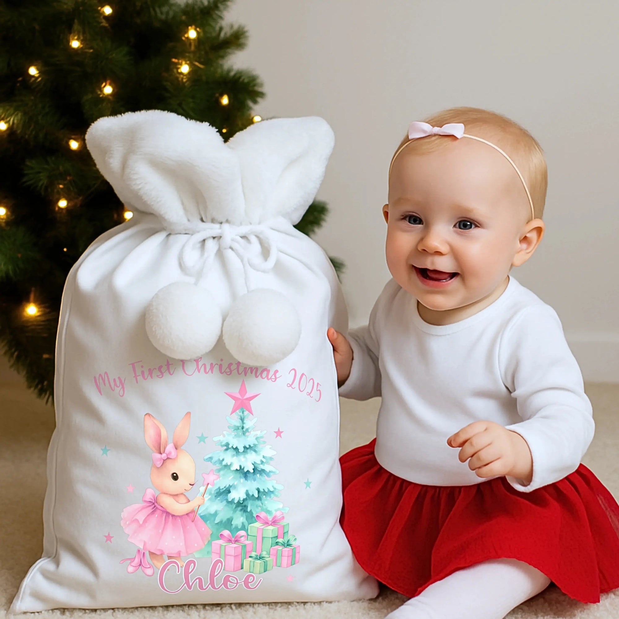 Smiling baby sitting beside a personalised Christmas Santa sack in front of a decorated Christmas tree, showcasing personalised Christmas gifts for children.