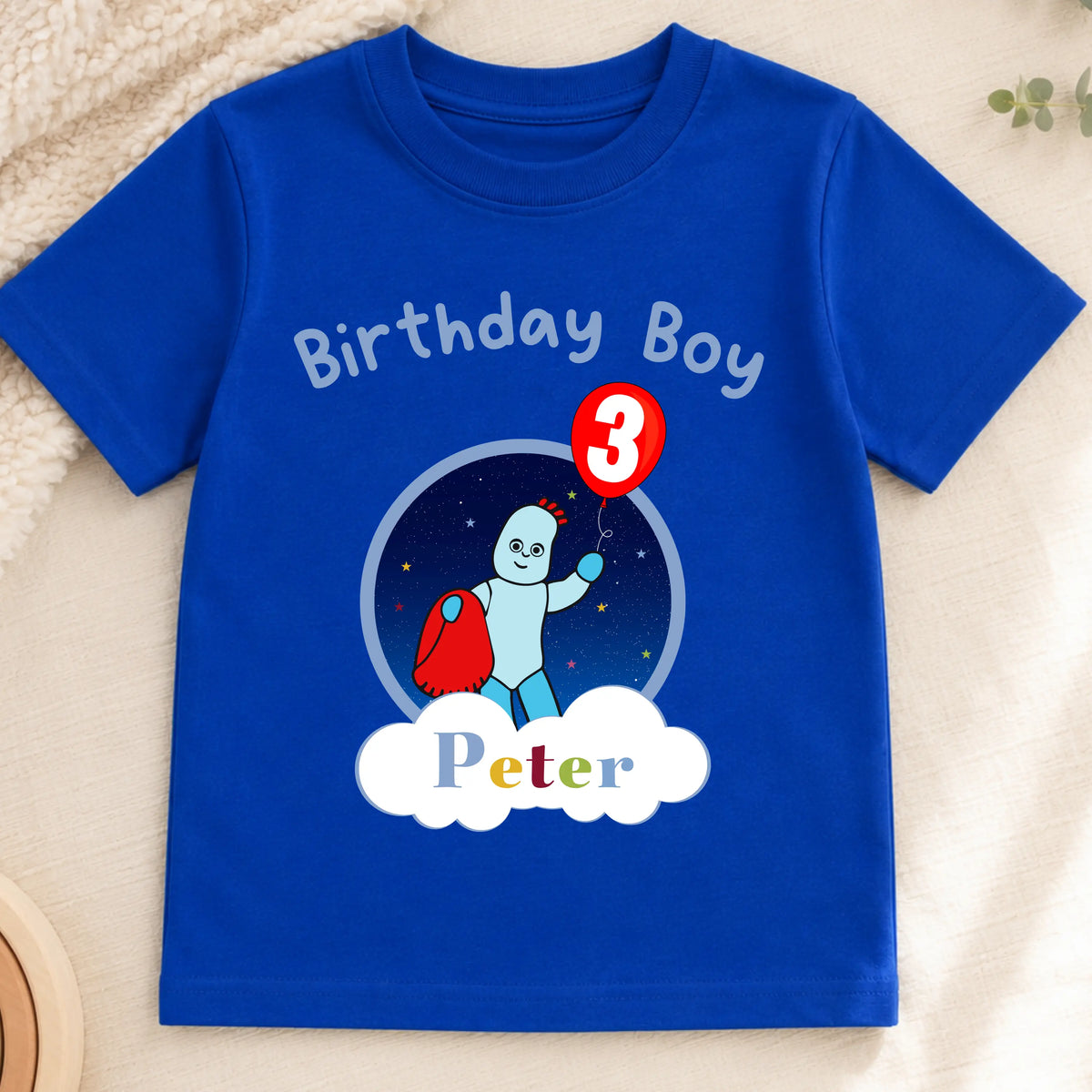 A personalised royal blue short-sleeve baby t-shirt with a 'Birthday Boy Peter' third birthday graphic, displayed on a neutral beige fabric background with wooden beads and a toy car. In the Night Garden theme with Iggle Piggle.