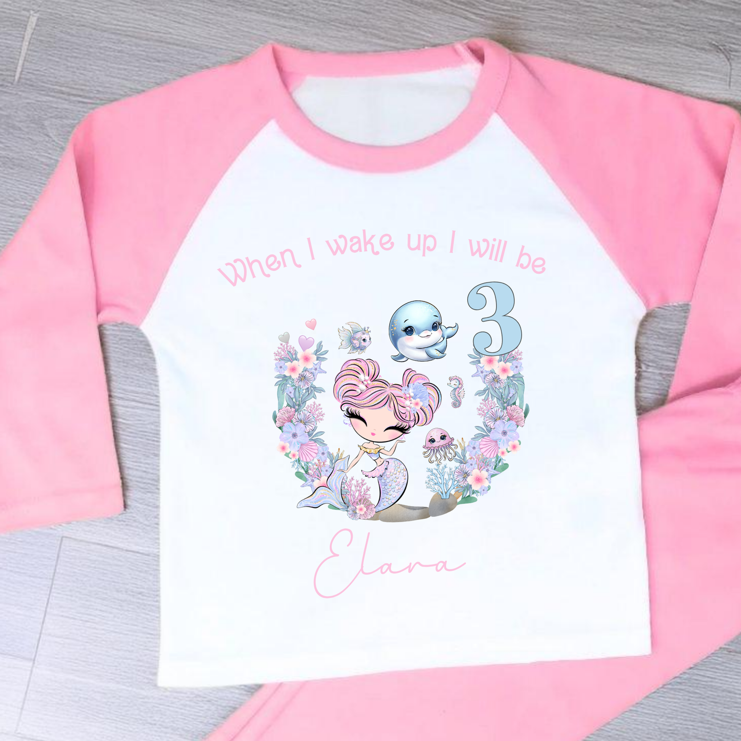 Mermaid birthday pyjamas, personalised for girls: A pair of mermaid birthday pyjamas in lilac and pink, featuring a glitter-like design. The top displays the phrase “When I Wake Up I Will be,” personalized with a girl's name and age. Ideal for young mermaid enthusiasts, these Pyjamas make a magical birthday gift for girls, when i wake up mermaid