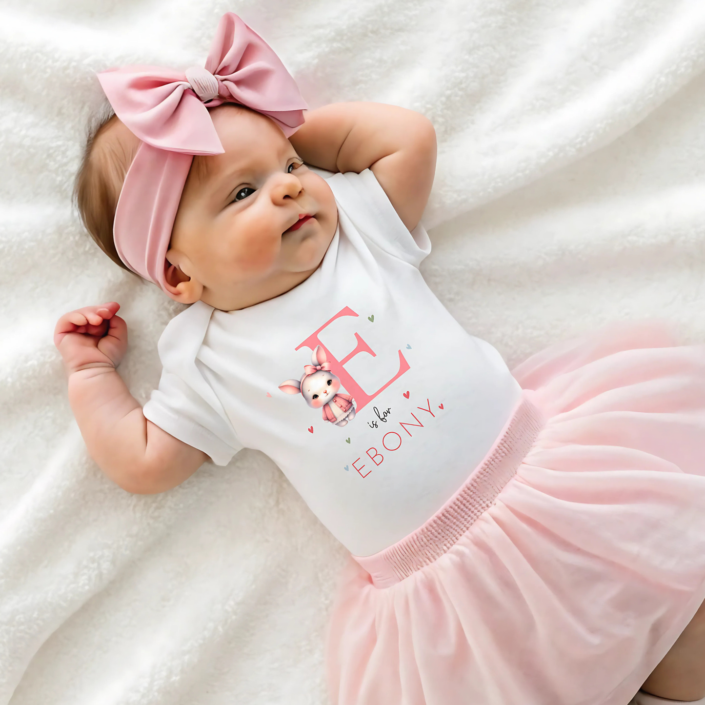 personalised baby outfit: An adorable baby girl wearing a pink sleepsuit for, featuring a cute dusky pink rabbit design. This personalised outfit is available as a sleepsuit, bodysuit, T-shirt, or bib, making it a perfect new baby gift. Ideal for coming home outfits or gender reveal announcements. It has the baby&#39;s initial and name along with the pink rabbit design.