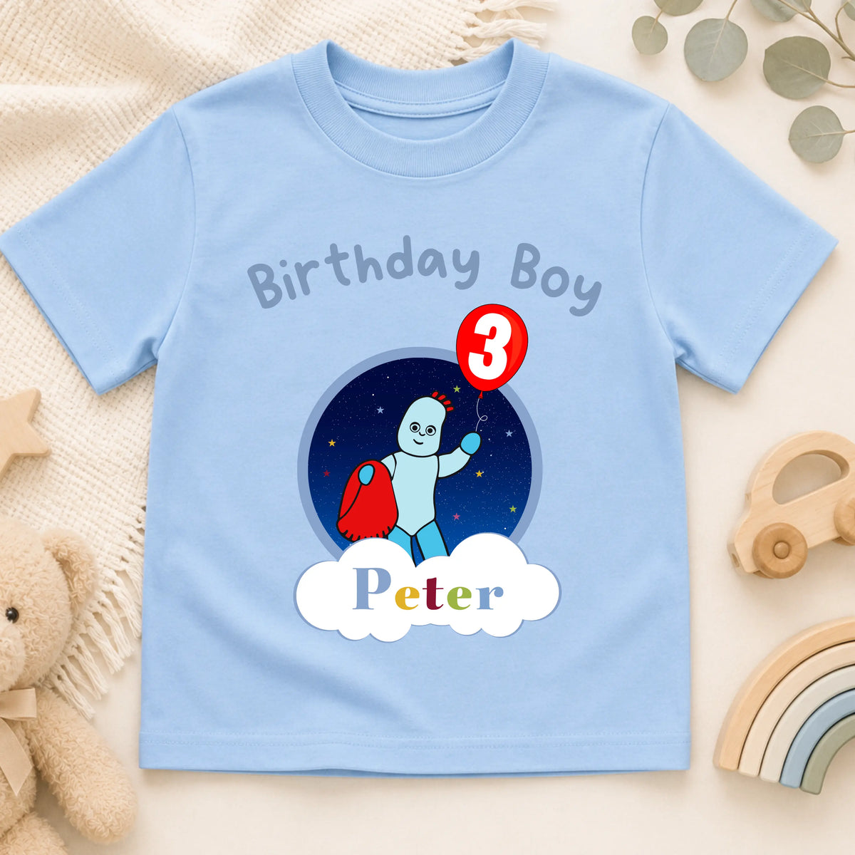 A personalised blue short-sleeve baby t-shirt with a 'Birthday Boy Peter' third birthday graphic, displayed on a neutral beige fabric background with wooden beads and a toy car. In the Night Garden theme with Iggle Piggle.