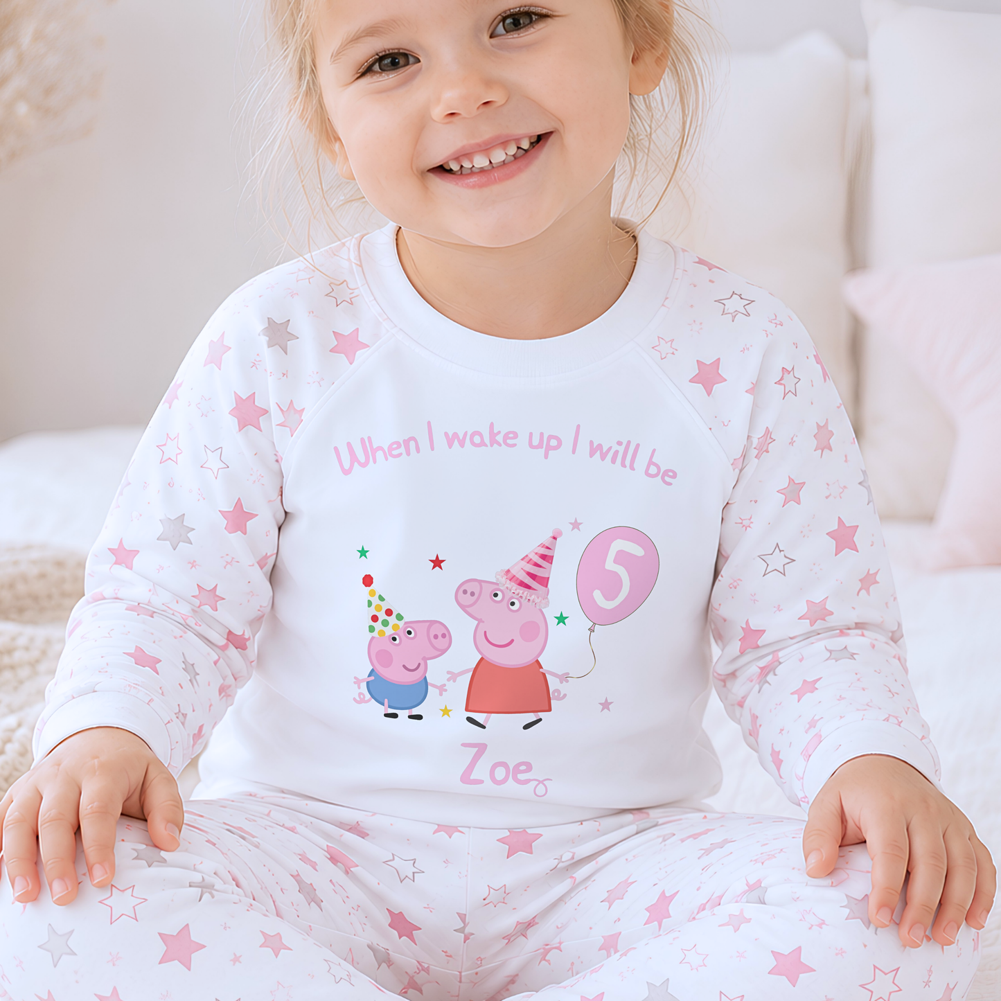 Girls Personalised Peppa and George Pig Birthday Pyjamas, A smiling young girl wearing a white long-sleeve pajama top and matching pants with pink star prints and a Peppa Pig birthday graphic on the front, sitting on a soft bed.