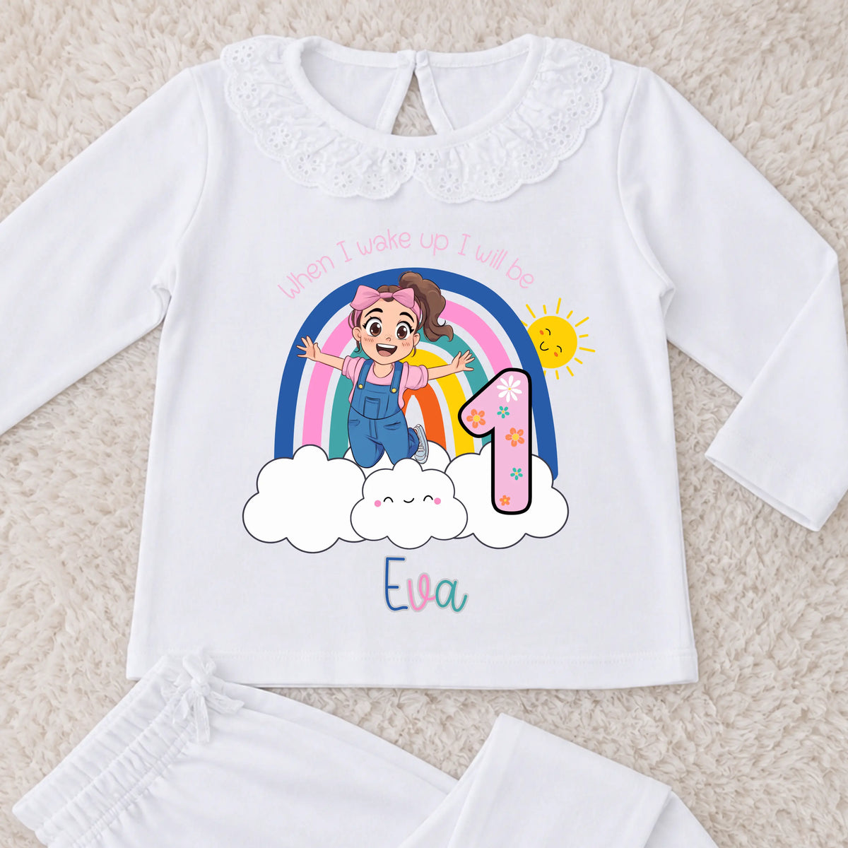 Personalised Ms Rachel Birthday PJs for Girls, When I Wake up Ms Rachel Pyjamas AishaBaileyDesigns: A white long-sleeve baby t-shirt with a lace collar, featuring a colorful first birthday graphic with a cartoon girl, rainbow, clouds, a number 1, and the name 'Eva'. White pants are partially visible at the bottom on a beige textured background.