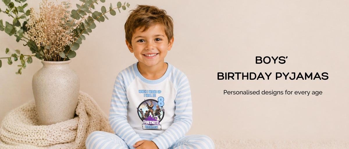 A smiling young boy wearing a light blue and white striped birthday pyjama set personaliseed on a cream rug in a styled room setting. To his left is a beige ceramic vase with dried botanicals and a knitted throw.