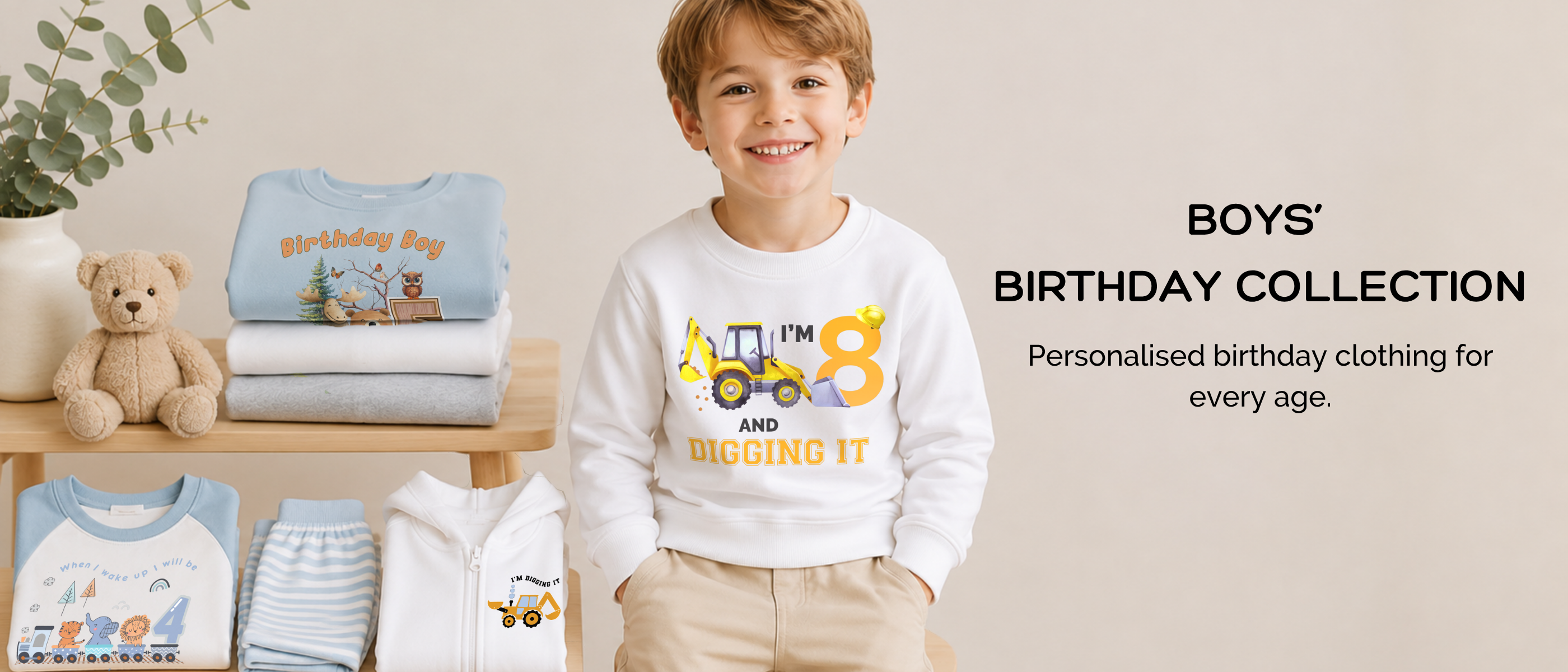 A smiling young boy stands indoors wearing a white long-sleeve birthday shirt with a yellow construction truck graphic and the words 'I'M 8 AND DIGGING IT.' To his left, folded matching baby clothes and a teddy bear sit on a wooden shelf, with text on the right reading 'BOYS' BIRTHDAY COLLECTION' and 'Personalised birthday clothing for every age.