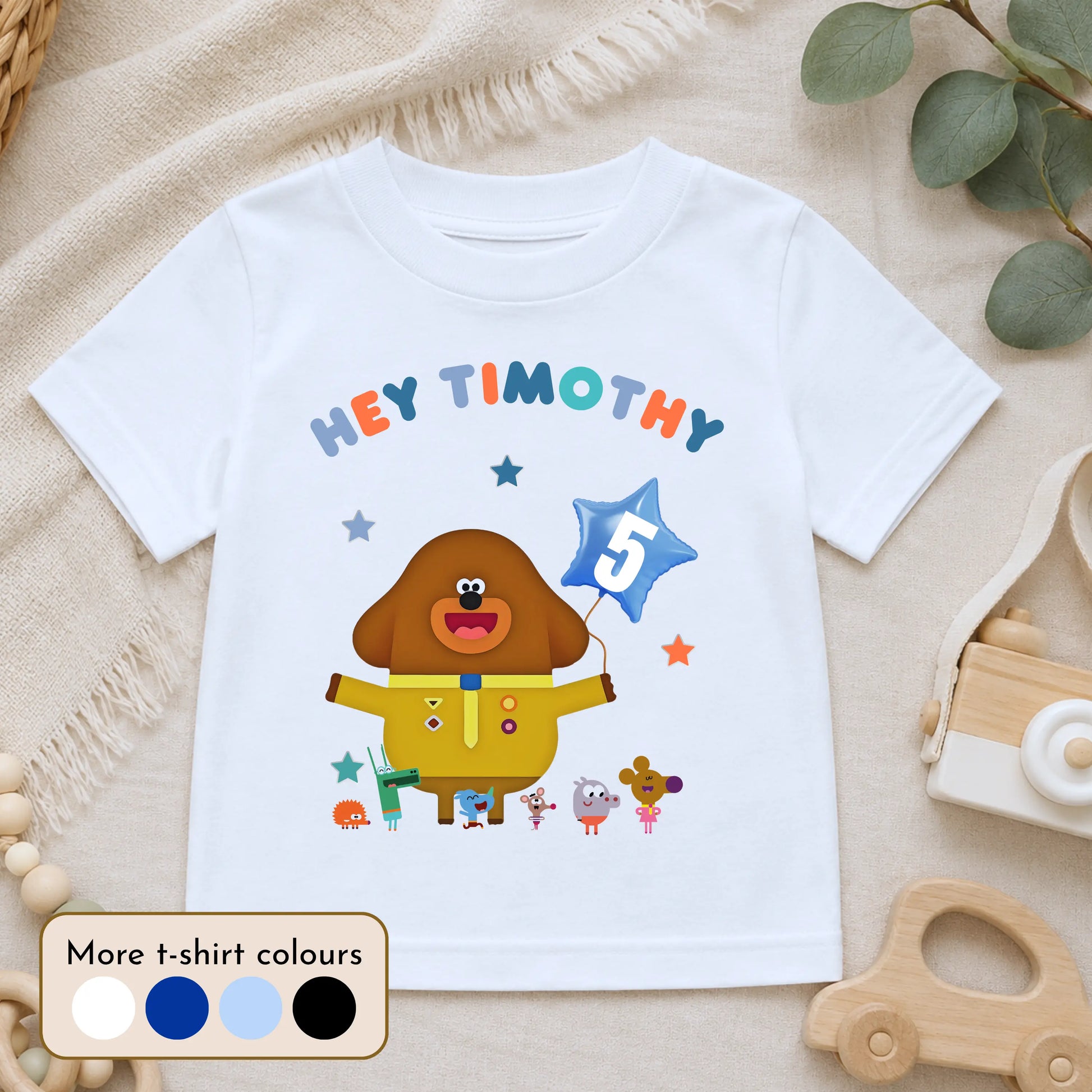 A  personalised white kids' t-shirt with a colorful Hey Duggee graphic and the text 'HEY TIMOTHY' on the front, styled in a flat-lay with a wooden toy car, eucalyptus leaves, and a cream fabric background.