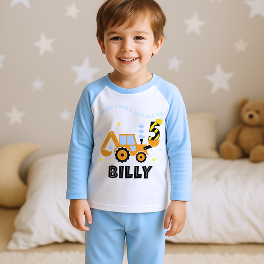 A boy wearing blue Pyjama set with a yellow excavator design. The excavator has a black bucket and a black age number on the side. The text 'When I Wake up I will be' is above the excavator and the child's name is below the excavator Digger Construction. Boys personalised birthday gift, Tractor birthday pyjamas, boys custom pyjamas with name, When I wake up construction digger, birthday pyjamas with name