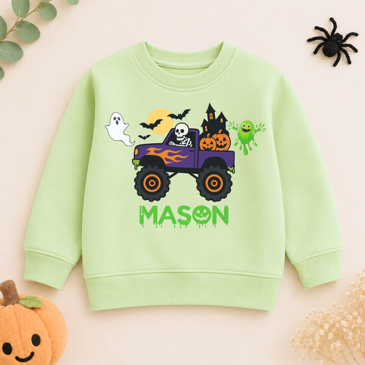 Personalised Halloween Monster Truck Sweatshirt, Halloween Monster Truck Sweatshirt with ghost, pumpkins, and personalized name for kids. A light green kids' sweatshirt with a colorful Halloween monster truck graphic and the name 'MASON' printed on the front, styled on a pale pink background with seasonal props.