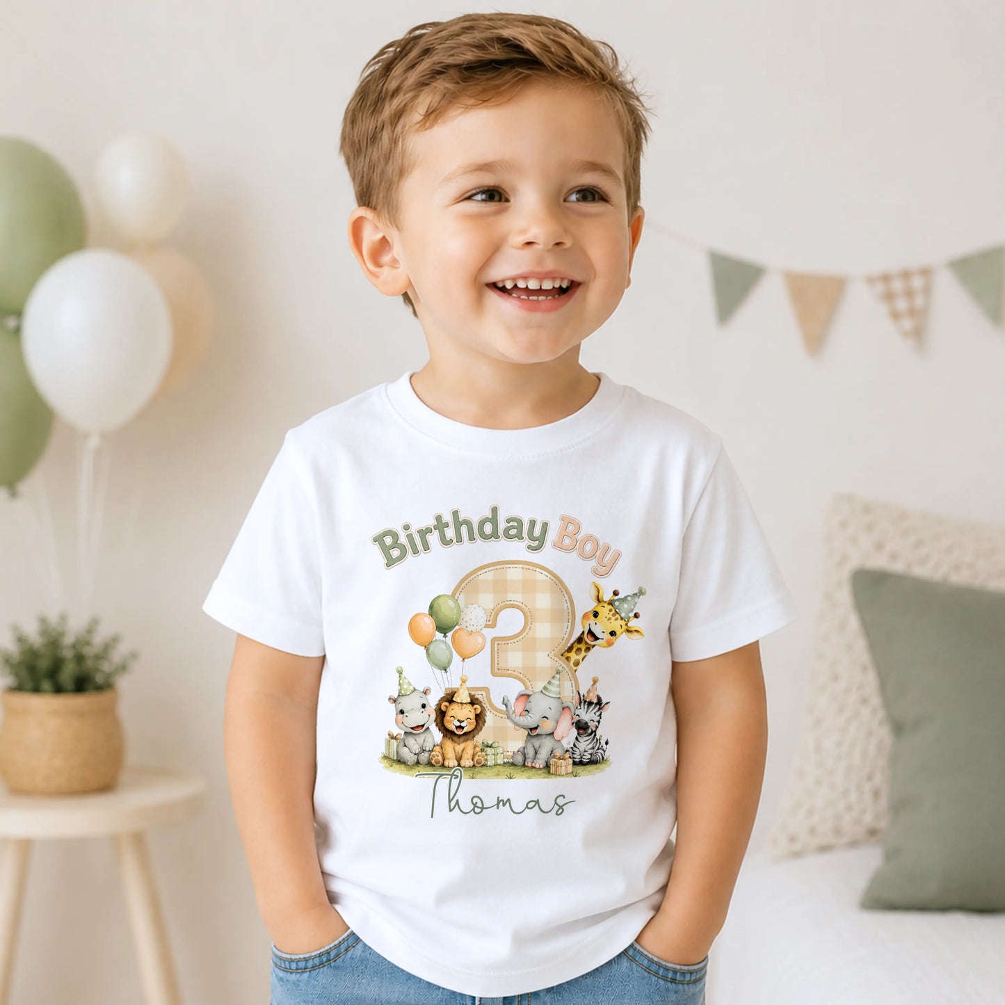 A smiling young boy wearing a white short-sleeve t-shirt with a colorful 'Birthday Boy Thomas' safari animal graphic, standing in a brightly lit, neutral-toned room.