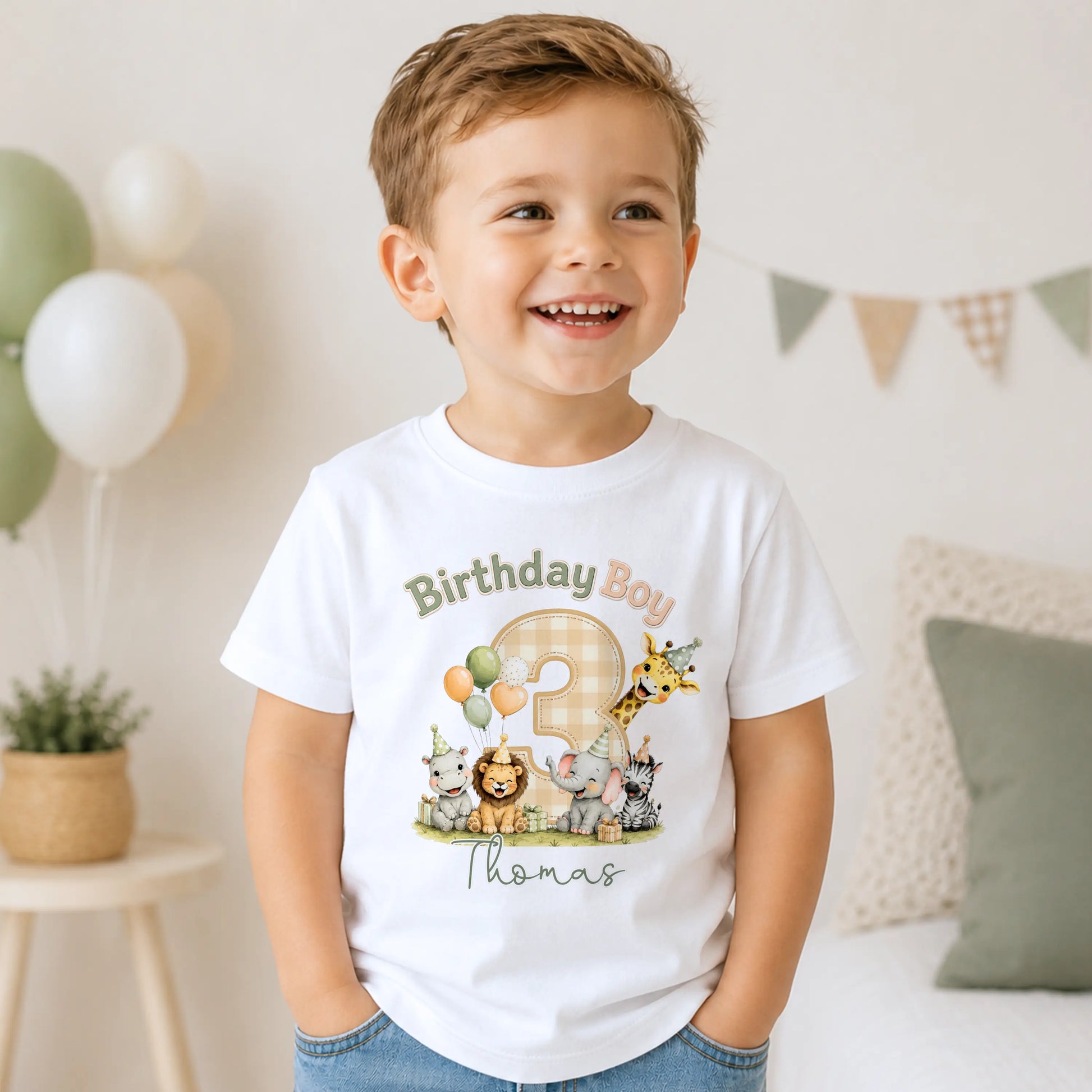 A smiling young boy wearing a white short-sleeve t-shirt with a colorful 'Birthday Boy Thomas' safari animal graphic, standing in a brightly lit, neutral-toned room.