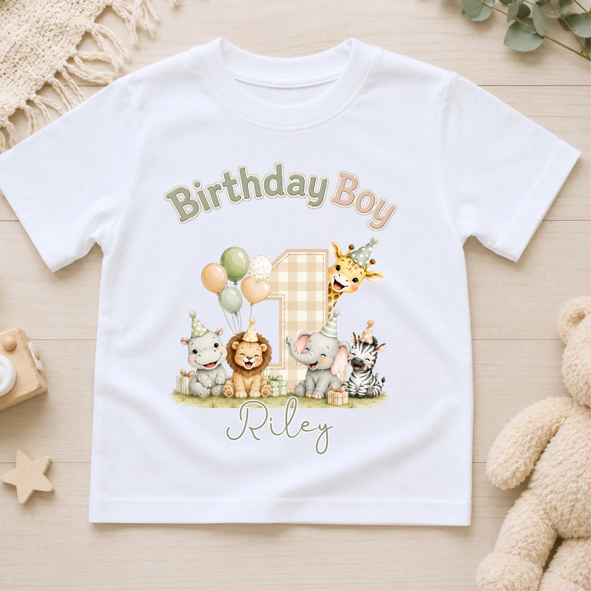 A white short-sleeve kids' t-shirt laid flat on a light wood surface, featuring a colorful safari-themed 'Birthday Boy' graphic with the name 'Riley' and an elephant, giraffe, lion, zebra, and cat illustration.