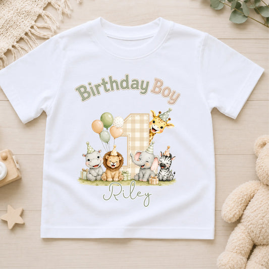 A white short-sleeve kids' t-shirt laid flat on a light wood surface, featuring a colorful safari-themed 'Birthday Boy' graphic with the name 'Riley' and an elephant, giraffe, lion, zebra, and cat illustration.