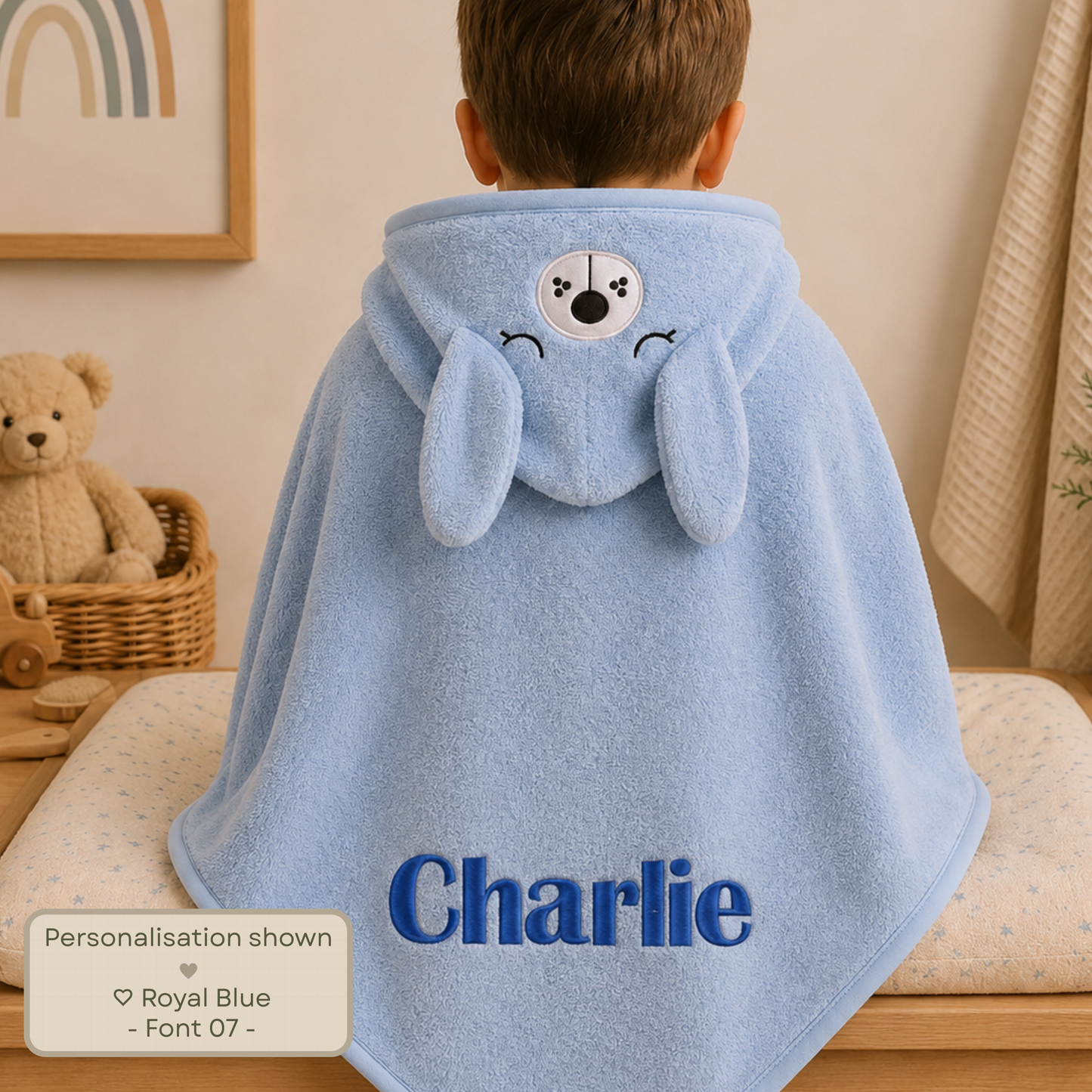 Personalised embroidered baby and toddler hooded towel with animal character hood, suitable from newborn to 3 years. A child seen from behind wearing a light blue personalized hooded towel cape with bunny ears and a dog face on the hood, in a softly styled nursery setting. The name 'Charlie' is printed in blue on the lower right.
