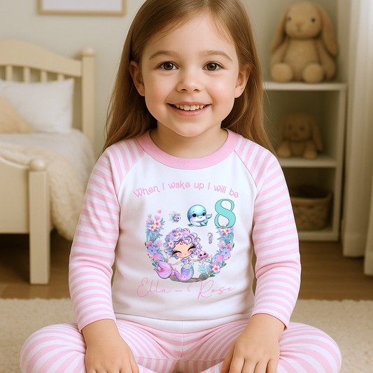 Mermaid birthday pyjamas, personalised for girls: A pair of mermaid birthday pyjamas in lilac and pink, featuring a glitter-like design. The top displays the phrase “When I Wake Up I Will be,” personalized with a girl's name and age. Ideal for young mermaid enthusiasts, these Pyjamas make a magical birthday gift for girls, when i wake up mermaid