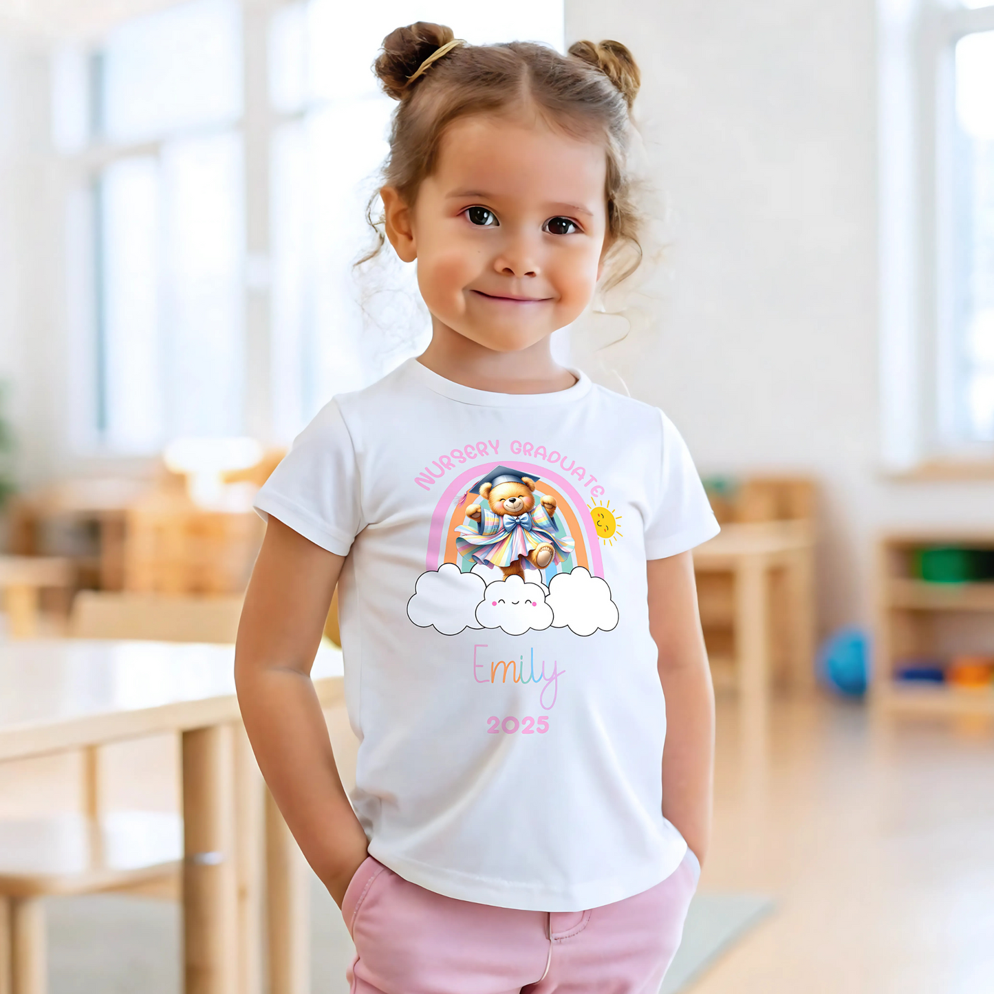 A girl wearing a white Personalised Nursery Leavers T-shirt. Above the rainbow it says Nursery Graduate and below it is a girl bear in graduation clothes on soft fluffy clouds. Below the design is the girls name and year of graduation.