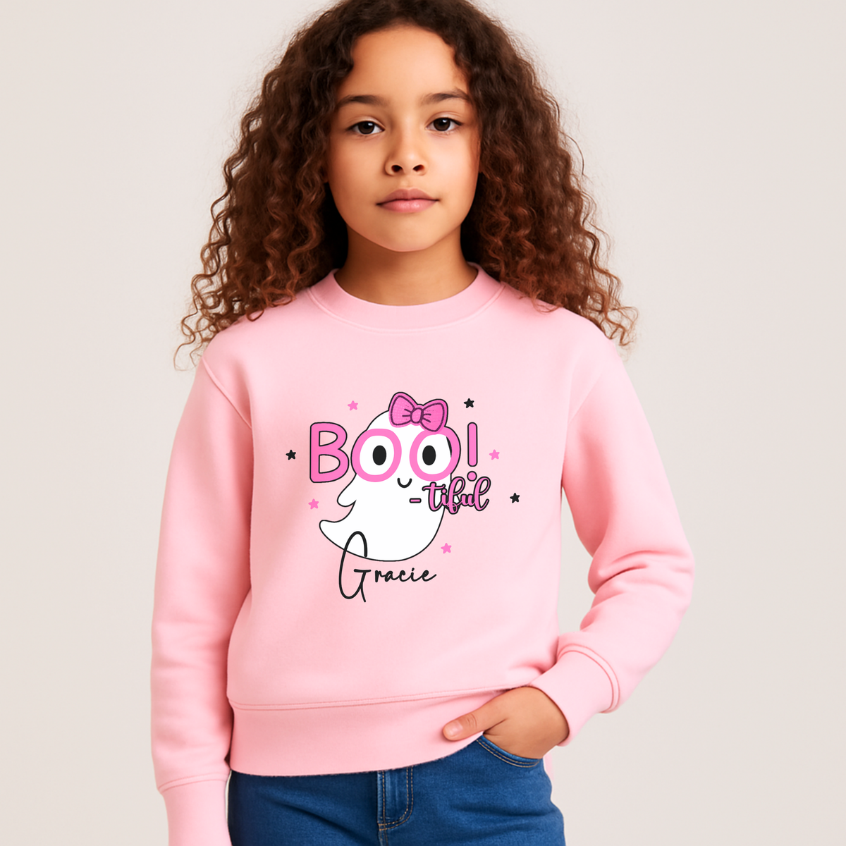 Personalised Girls Halloween Sweatshirt - Personalised Halloween sweatshirt for girls with cute ghost “Boo” design in lilac. A young girl with curly brown hair wearing a light pink crewneck sweatshirt with a 'BOO! that's Gracie' ghost graphic, paired with blue jeans, against a solid light gray background.