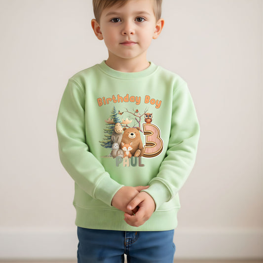 Woodland animal birthday sweatshirt: A children's birthday jumper featuring a woodland animal theme. The shirt displays a forest scene with a playful fox, a curious bear, and a gentle bear, surrounded by lush greenery. The design uses earthy tones and vibrant greens to create a natural look. At the top, the words “Birthday Boy” are printed, with space for a personalized name underneath. 