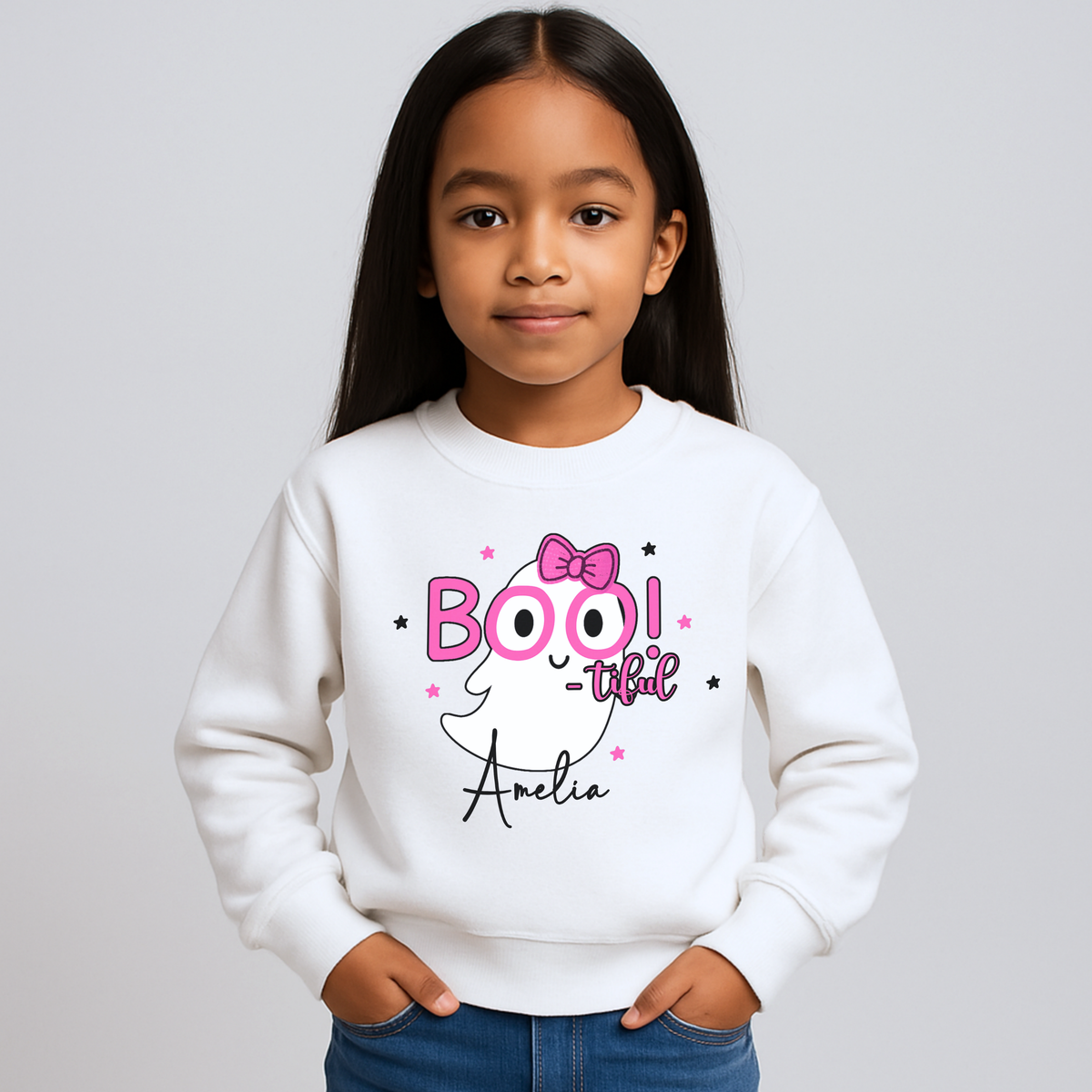 Personalised Girls Halloween Sweatshirt - Personalised Halloween sweatshirt for girls with cute ghost “Boo” design in lilac. A young girl with long dark hair wearing a white crewneck sweatshirt with a 'BOO! -that- Amelia' graphic on the front, paired with blue jeans, against a light gray studio background.