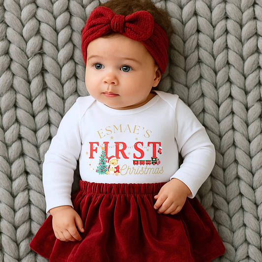 First Chrstmas Outfit, Baby wearing a white long-sleeve shirt with personalised  First Christmas' text and a red skirt, lying on a textured gray blanket.