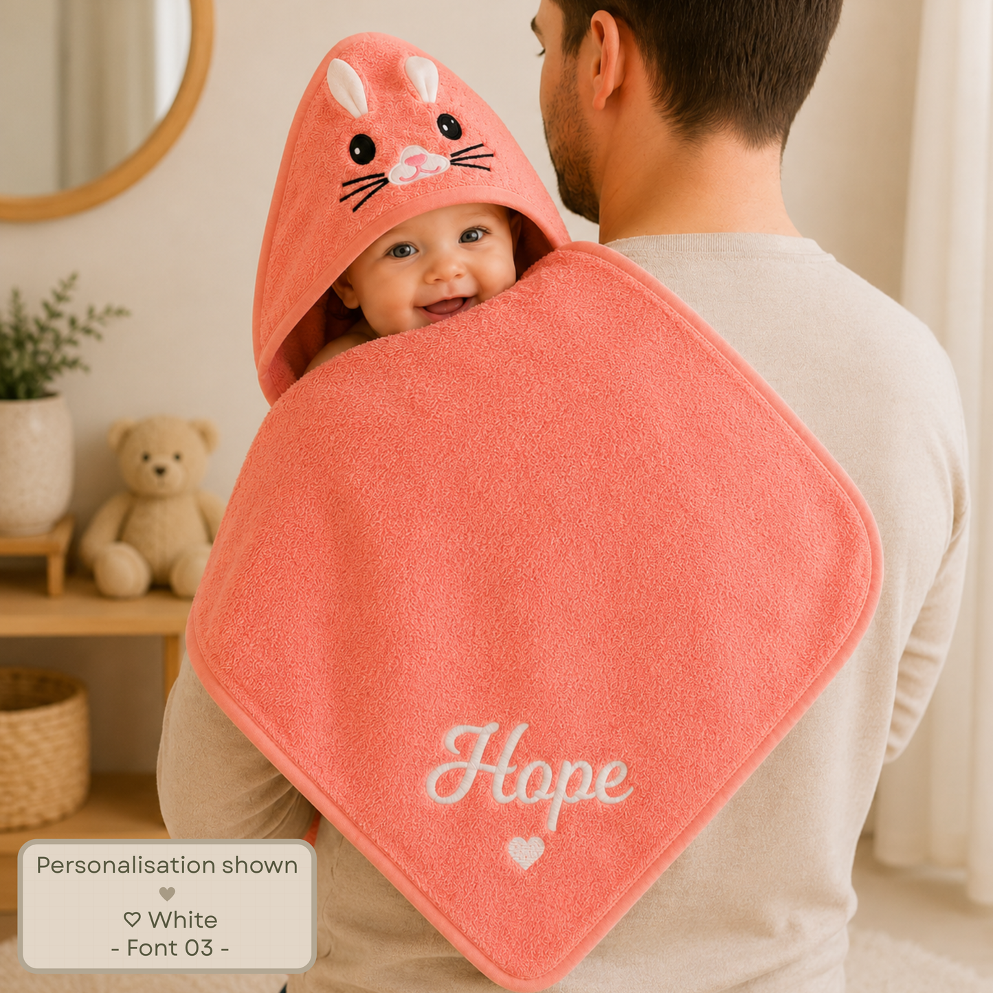 Personalised embroidered baby and toddler hooded towel with animal character hood, suitable from newborn to 3 years. A baby wrapped in a large peach-colored hooded towel with a bunny face on the hood, held by an adult in a bathroom setting. The towel has the name 'Hope' embroidered in white script at the bottom.