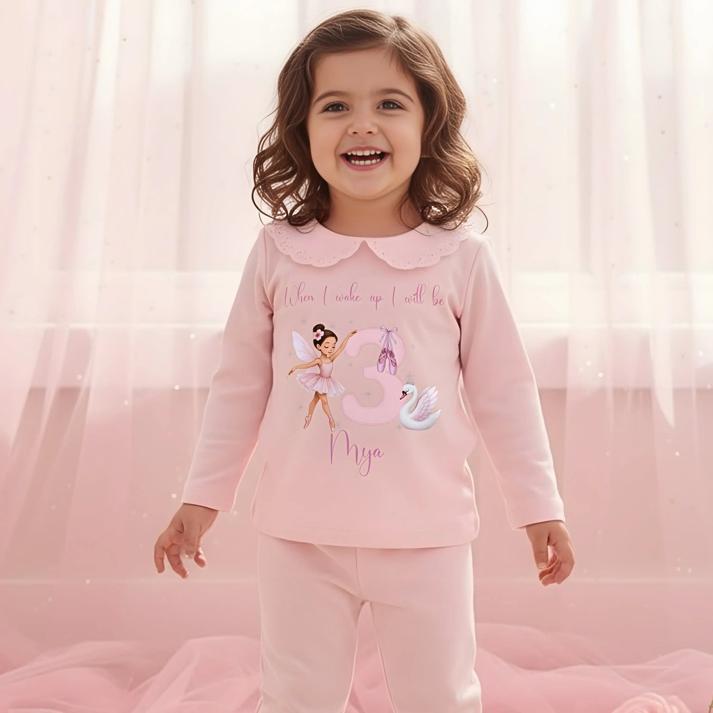 Ballerina Birthday Pyjamas - Young girl wearing a pink outfit with a fairy design and text, sitting on a carpeted floor.