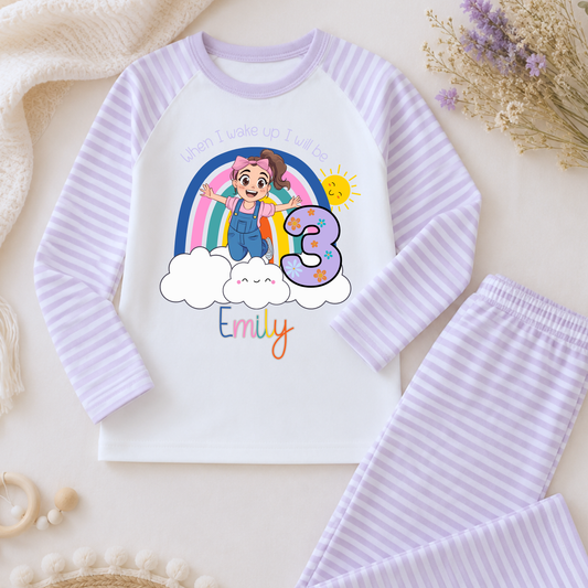 Personalised Ms Rachel Birthday PJs for Girls, When I Wake up Ms Rachel Pyjamas AishaBaileyDesigns: A children's two-piece pajama set laid flat on a light cream background with soft decorative props. The long-sleeve top is white with light lavender stripes on the sleeves and collar, featuring a colorful cartoon girl under a rainbow, a large number 3, and the name Emily.