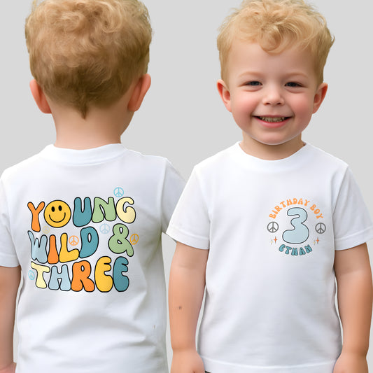 Personalised Young Wild and three birthday t-shirt: A boys birthday T-shirt with a Young Wild and Three theme, showcasing a groovy retro style. The front features whimsical retro emojis and the words birthday boy alongside a custom name on the left breast. The back displays bold retro fonts and graphics. Tthis vibrant and playful tee is designed for comfort and style, perfect for third birthday gifts.