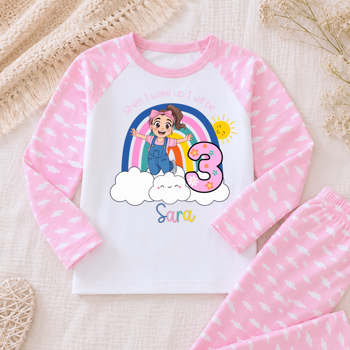 Personalised Ms Rachel Birthday PJs for Girls, When I Wake up Ms Rachel Pyjamas AishaBaileyDesigns:A children's two-piece pajama set laid flat on a soft beige fabric background. The long-sleeve top is white with pink sleeves and pink leaf patterns, featuring a colorful cartoon birthday graphic with the text 'when I wake up I feel.