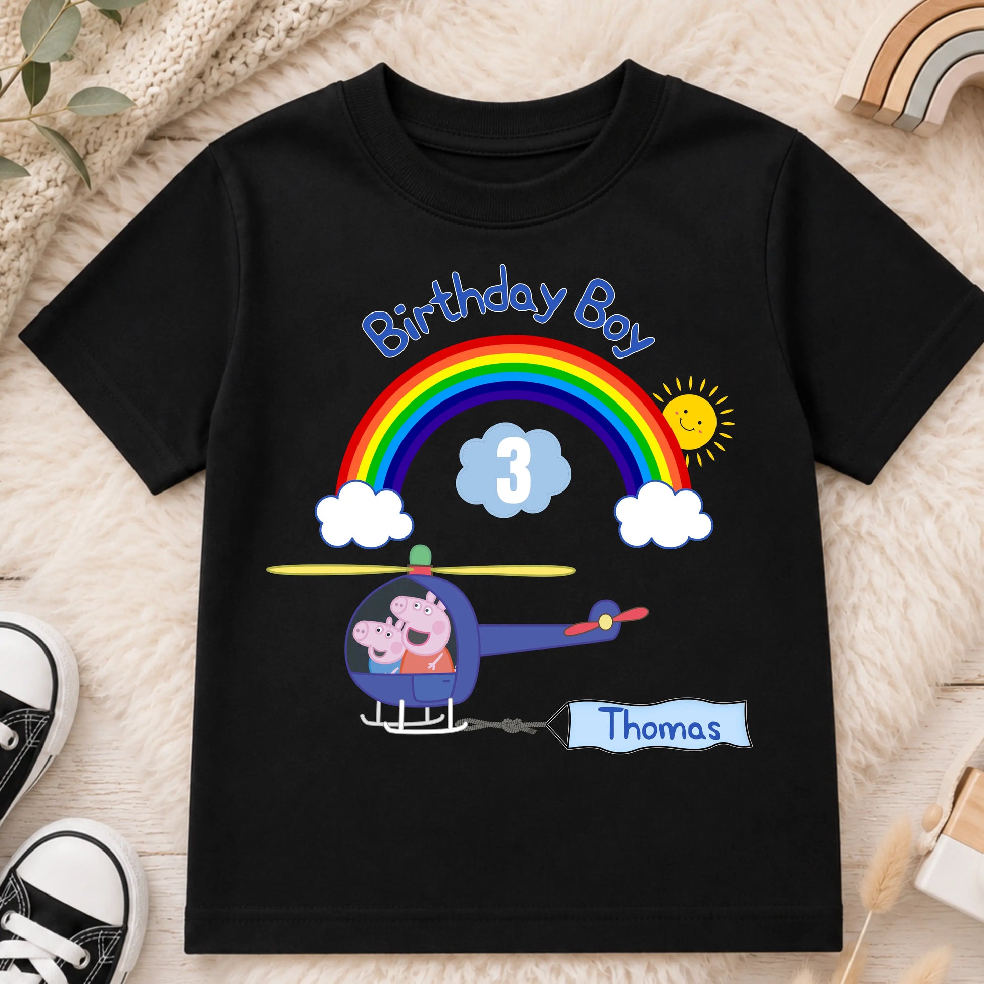 A black kids' t-shirt laid flat on a cream faux fur surface, featuring Personalised Peppa Pig and George birthday T-shirt for boys featuring a helicopter, rainbow, custom name Thomas and age 3 birthday design.