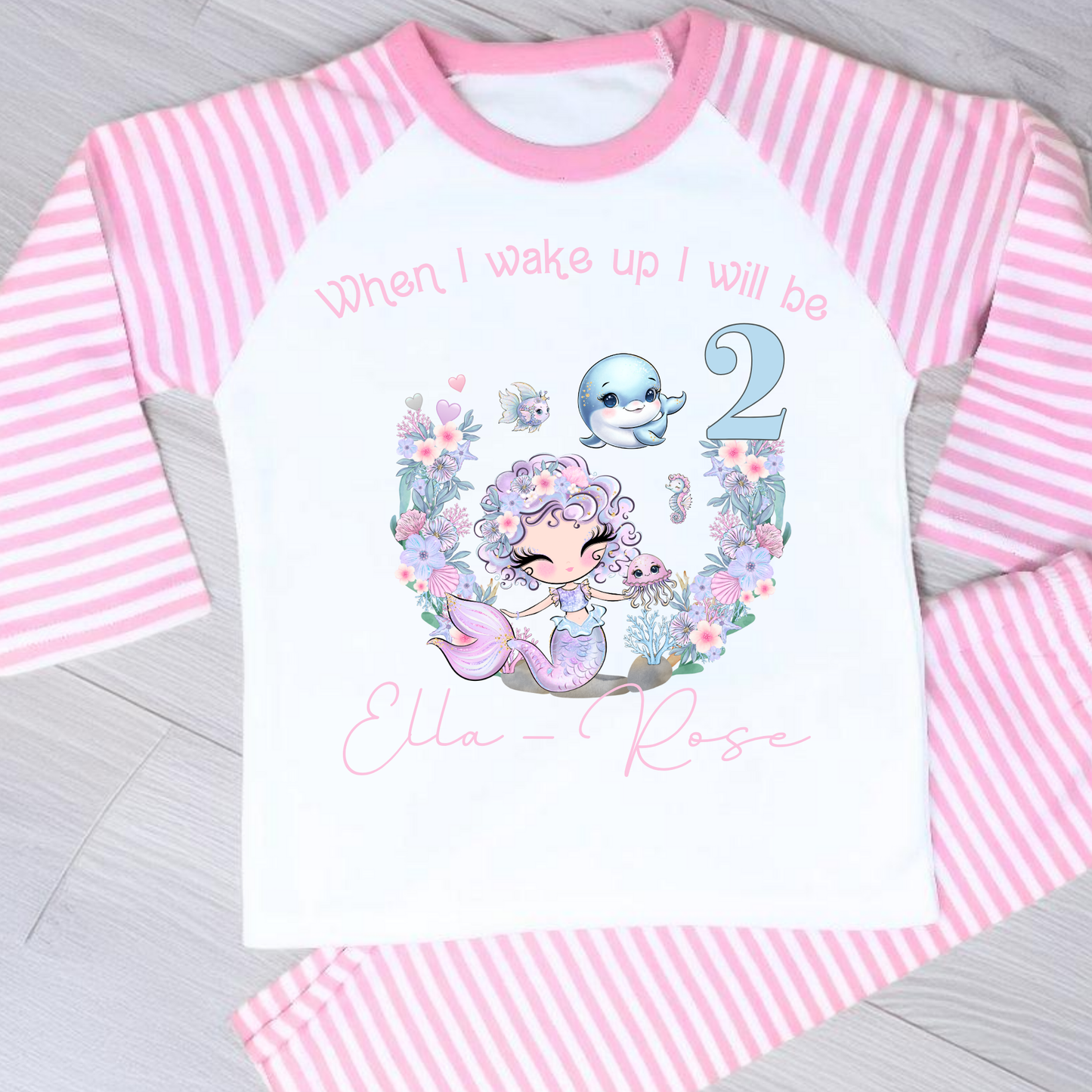 Mermaid birthday pyjamas, personalised for girls: A pair of mermaid birthday pyjamas in lilac and pink, featuring a glitter-like design. The top displays the phrase “When I Wake Up I Will be,” personalized with a girl's name and age. Ideal for young mermaid enthusiasts, these Pyjamas make a magical birthday gift for girls, when i wake up mermaid